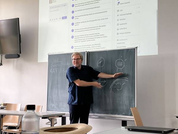 Ein Mann in dunkelblauer Kleidung und grauen Haaren lächelt in die Kamera und erklärt den Teilnehmenden eines Seminars etwas an der Tafel, über der Tafel erscheint die digitale Präsentation.